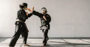 Full-body shot of an instructor and a student in black uniforms practicing striking and blocking movements in a spacious studio during self defense training for beginners.