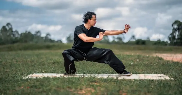 A man in a black shirt and loose pants practices a low, wide side-lunge stance on a mat outdoors, showcasing a martial arts posture from Tai Chi for Beginners.