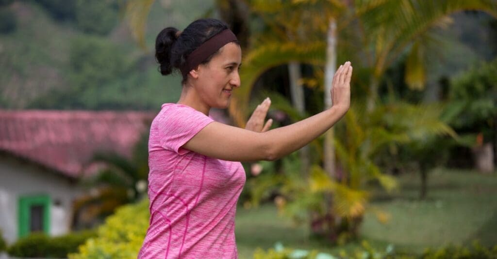 Close-up profile of a woman with a headband in a pink shirt practicing a slow, meditative hand gesture outdoors, illustrating a gentle movement from Tai Chi for Beginners.