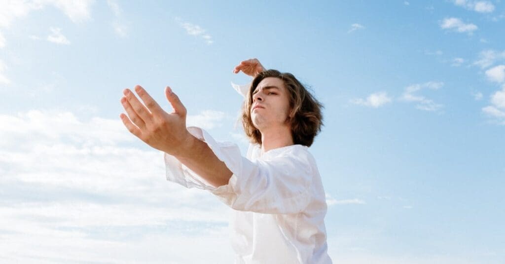 A man with long hair in a white shirt stands against a blue sky, raising one hand with open palms as part of an exercise from Tai Chi for Beginners.