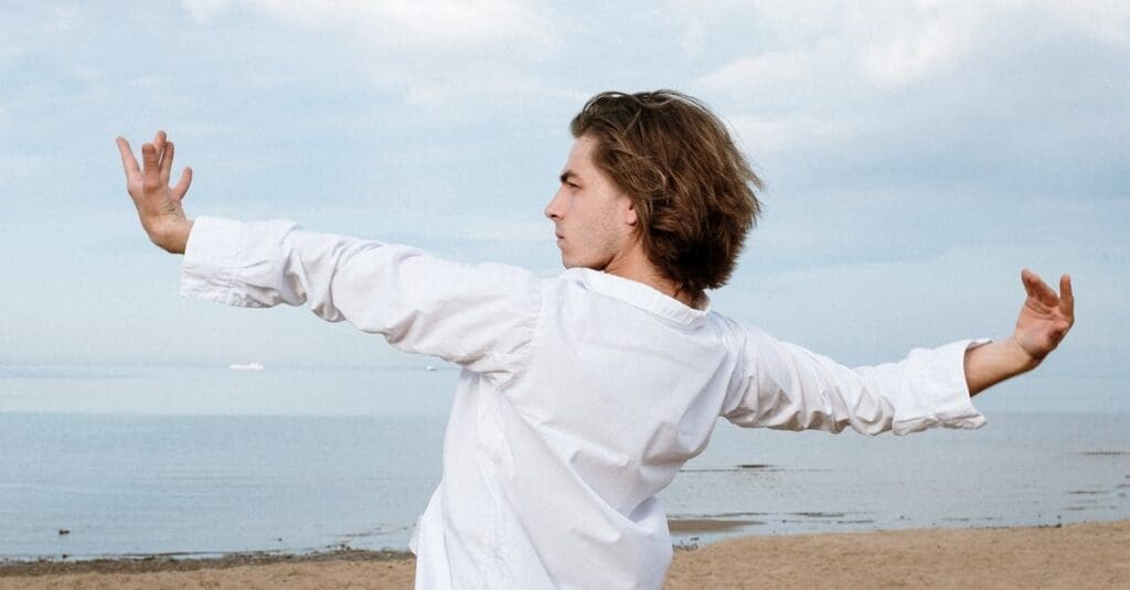 A man with long hair wearing a white shirt extends both arms outward while standing on a beach, practicing a balancing posture from Tai Chi for Beginners.