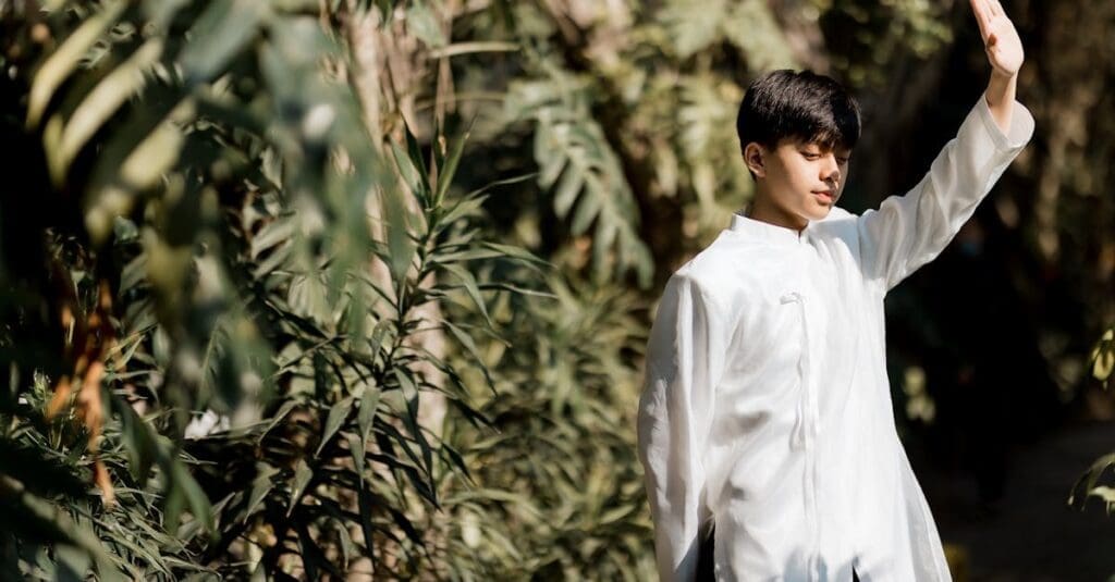 A young man in a white martial arts uniform raises his hand in a slow, controlled motion among lush green foliage, demonstrating a pose from Tai Chi for Beginners.
