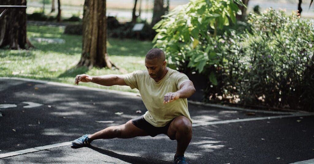A man in athletic wear performs a deep side lunge with extended arms on an outdoor paved path, illustrating a flexibility routine from Tai Chi for Beginners.