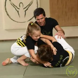 Young girl practicing a ground control technique with a partner while an instructor provides guidance during a kids martial arts class focused on ground fighting in Brazilian Jiu Jitsu.