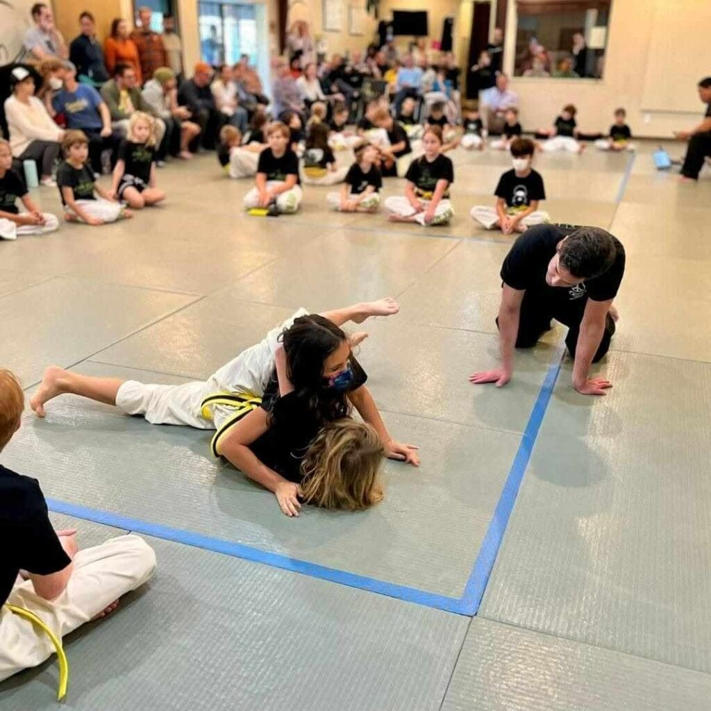 Two children practicing a pinning and escape drill on the mat while classmates watch during a lesson on ground fighting in Brazilian Jiu Jitsu.